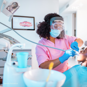 A dental care provider wearing protective equipment, including a face shield, mask, and gloves, treats a child patient in a dental exam room. Dental tools and a monitor displaying teeth imagery are visible in the PHC-branded pediatric dental setting.