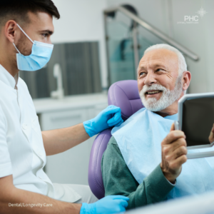 An older adult patient sitting in a dental chair smiles while holding a mirror and speaking with a dental care provider wearing a mask and gloves. The PHC-branded image represents dental and longevity care in a modern clinic setting.