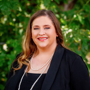 Portrait of Stephany Balow, smiling and wearing a black blazer, layered pearl necklaces, and a red lip, with a green leafy background.