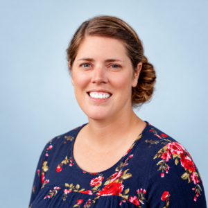 Portrait of Julie Raasch, smiling and wearing a floral-patterned navy blouse, with a green leafy background.