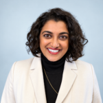 Dr. Megan Srinivas, Chief Medical Officer at Primary Health Care, smiling in a professional headshot wearing a tan blazer and black top against a blue background